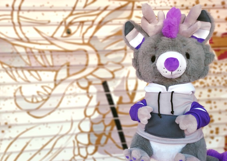 May include: A gray plush toy with purple accents, wearing a gray hoodie with purple accents. The toy has a purple nose, purple eyes, and a purple mohawk. The toy is sitting in front of a white and brown background.