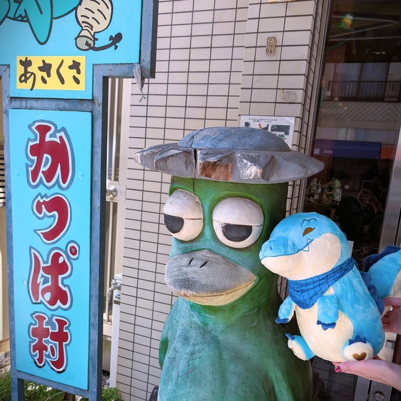 May include: A green wooden statue of a kappa, a Japanese folkloric creature, wearing a hat. A blue plush toy of a crocodile is held next to the statue. A blue sign with Japanese text is on the left.
