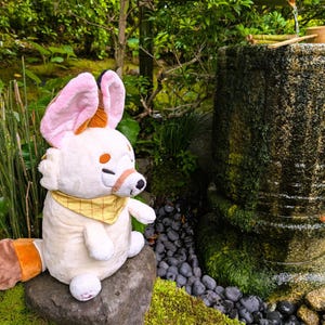 May include: A plush toy fox with large pink and brown ears, a yellow bandana, and a white body sits on a rock. The fox is in a garden setting with a stone water basin and lush greenery in the background.