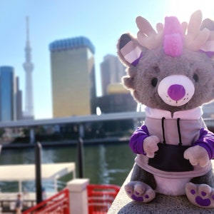 May include: A gray plush toy with purple and white antlers, wearing a gray hoodie with purple sleeves. The toy is sitting on a concrete surface with a blurred city skyline in the background.