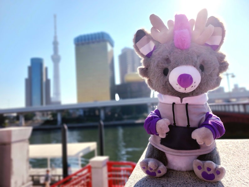 May include: A gray plush toy with purple and white antlers, wearing a gray hoodie with purple sleeves. The toy is sitting on a concrete surface with a blurred city skyline in the background.