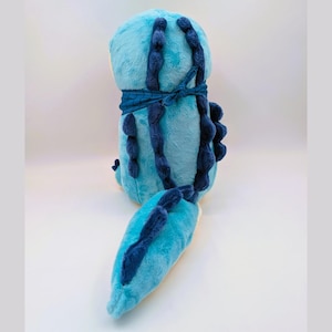 May include: A plush toy in shades of blue, featuring a rounded body and a long tail. Dark blue accents include a band around the middle and decorative elements along the back and tail. The toy is soft and cuddly.