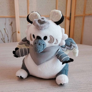 May include: A plush toy griffin with a white body, gray and blue accents, and a gray bandana. The griffin has large, round eyes, a blue beak, and detailed wings. The toy is sitting on a light-colored surface.