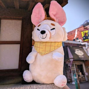 May include: A plush toy in the shape of a smiling, cream-colored animal with large pink ears. It has a yellow checkered bandana around its neck and small pink paw pads. The toy is sitting on a ledge.