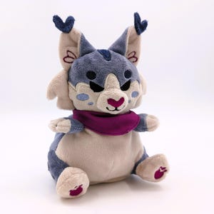 Puede incluir: Un peluche de lince con una bufanda morada. El peluche tiene un cuerpo beige claro, detalles azules y grises y una nariz rosa. El lince tiene orejas grandes con puntas azul oscuro y pequeños detalles bordados.