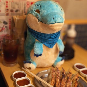 May include: A plush blue and tan alligator toy with a blue bandana. The stuffed animal is sitting on a wooden table. A glass of iced tea, dipping sauces, and a plate of food are also on the table.