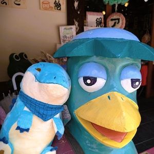 May include: A whimsical, colorful display featuring two cartoonish sculptures. One is a blue dinosaur with a blue scarf, and the other is a teal-colored duck-like figure with a yellow beak and a hat. The sculptures are painted with bright colors.