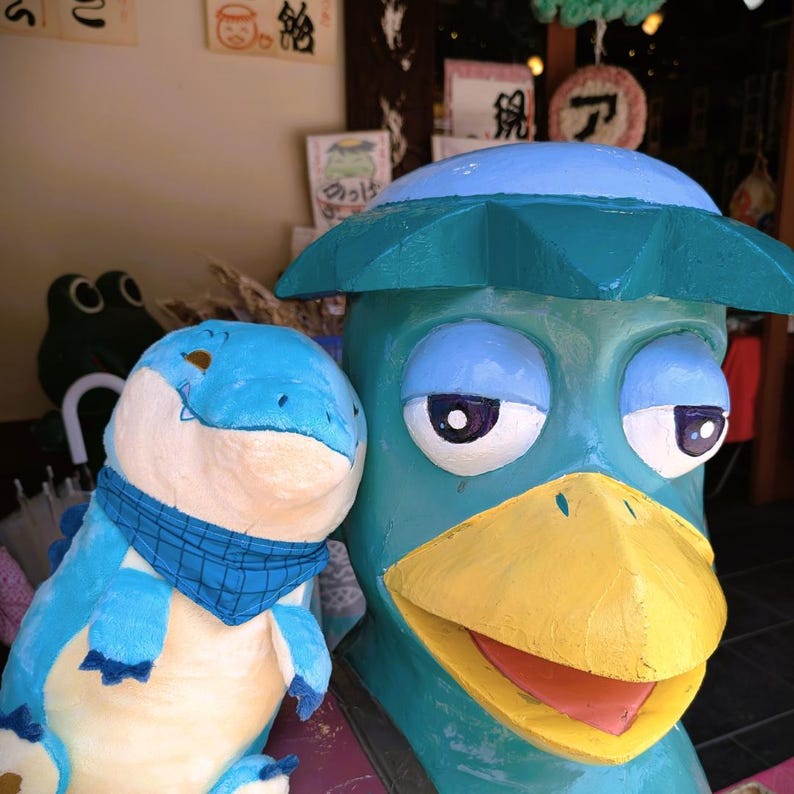 May include: A whimsical, colorful display featuring two cartoonish sculptures. One is a blue dinosaur with a blue scarf, and the other is a teal-colored duck-like figure with a yellow beak and a hat. The sculptures are painted with bright colors.