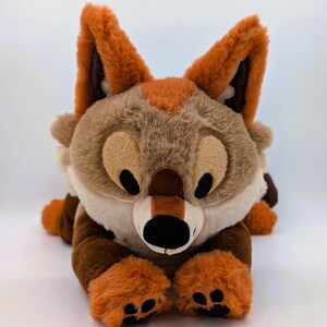Puede incluir: Un peluche de zorro en tonos marr&oacute;n, beige y naranja, tumbado sobre una superficie blanca. El zorro tiene ojos grandes y expresivos, una nariz negra y patas naranjas. Las orejas son de un marr&oacute;n m&aacute;s oscuro con puntas naranjas.
