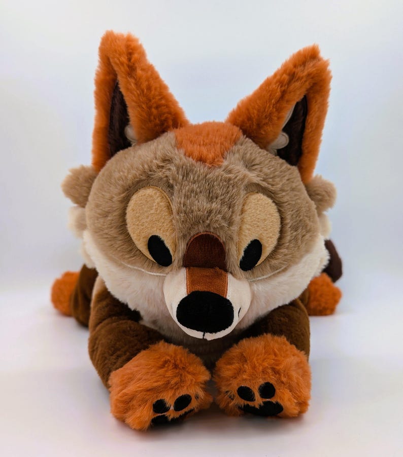 Puede incluir: Un peluche de zorro en tonos marr&oacute;n, beige y naranja, tumbado sobre una superficie blanca. El zorro tiene ojos grandes y expresivos, una nariz negra y patas naranjas. Las orejas son de un marr&oacute;n m&aacute;s oscuro con puntas naranjas.