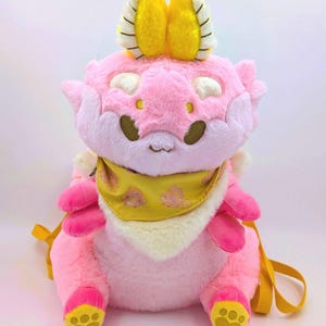 Puede incluir: Una mochila de peluche rosa con forma de criatura de dibujos animados. Tiene detalles amarillos en la cabeza y las patas, y un pañuelo amarillo. La mochila tiene correas amarillas y una cara sonriente.