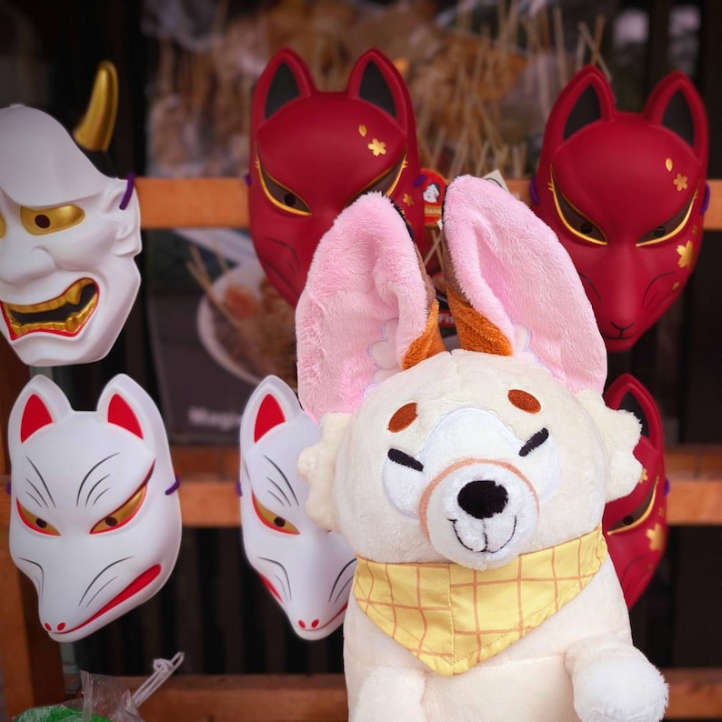 May include: A display of Japanese fox and demon masks, alongside a plush toy with large pink ears and a yellow scarf. The masks are red and white with gold accents. The plush toy is cream-colored.