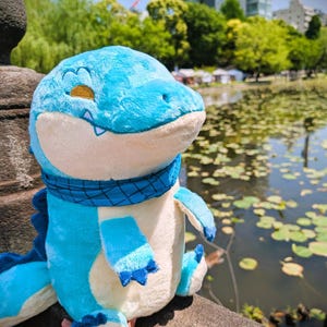May include: A plush blue dinosaur toy with a cream-colored belly and a blue scarf. The dinosaur has a friendly expression with closed eyes and a stitched smile. The background shows a pond with lily pads and trees.