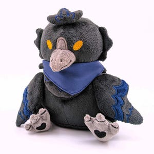 May include: A plush crow toy with a dark grey body, a grey beak, and orange eyes. It wears a blue bandana and a small hat with blue embroidery. The crow's wings and feet have blue accents and black heart details.
