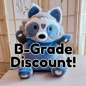 Może przedstawiać: Pluszowy szop w kolorze szarym i niebieskim z niebieskim szalikiem i napisem "B-Grade Discount!" czarnymi literami.