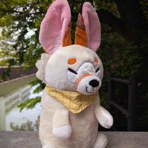 May include: A plush toy animal with large pink and brown ears, orange eyes, and a yellow bandana. The stuffed animal has a cream-colored body and is sitting upright. The toy has a smiling expression.