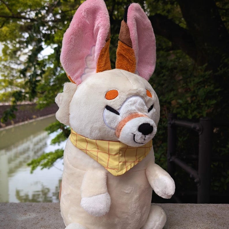 May include: A plush toy animal with large pink and brown ears, orange eyes, and a yellow bandana. The stuffed animal has a cream-colored body and is sitting upright. The toy has a smiling expression.