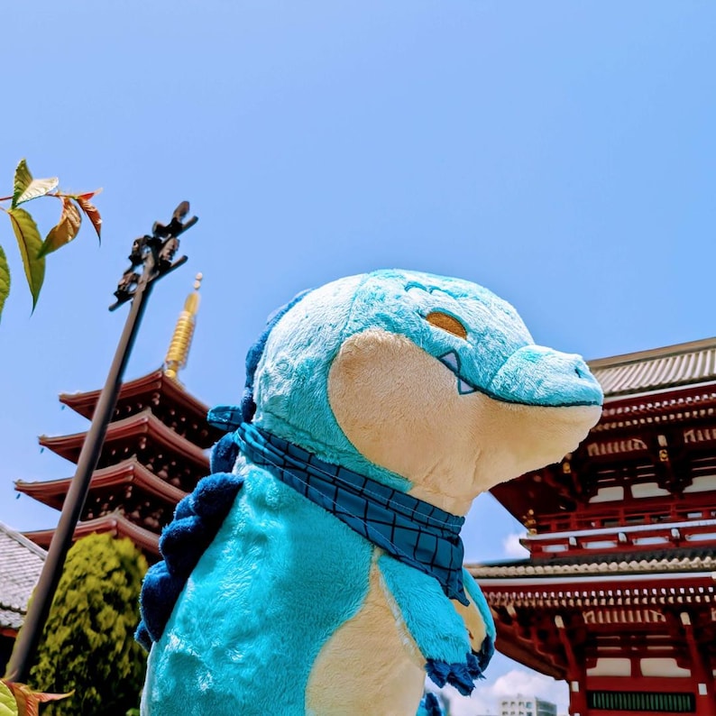 May include: A plush, light blue crocodile toy with a cream-colored snout and yellow eyes. It wears a dark blue scarf and has dark blue spines down its back. The background features a pagoda and red-roofed buildings under a clear, blue sky.