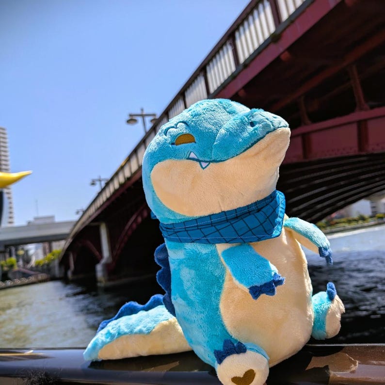 May include: A plush alligator toy in shades of blue and cream, wearing a blue patterned scarf. The toy has a smiling expression and is positioned outdoors with a bridge and water in the background. The alligator's tail and feet are visible.