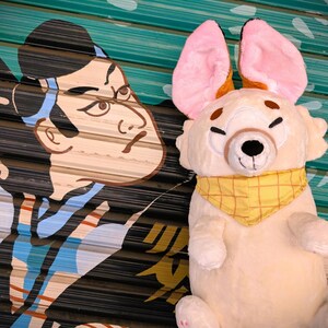 May include: A plush toy dog with large pink ears and a yellow bandana. The dog has a cream-colored body, black eyes, and brown accents. The background features a mural of a Japanese figure.