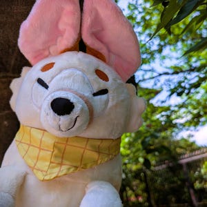 May include: A plush toy in the shape of a white bunny with large pink ears. The bunny has a black nose, closed eyes, and a yellow bandana with a plaid pattern. The background is blurred with green foliage.