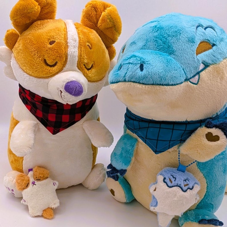 May include: Two plush toys: a corgi-like dog with orange and white fur, wearing a red and black plaid bandana, and a blue alligator-like creature with a blue bandana. A small plush dog is in front of the corgi.