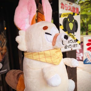 May include: A plush toy in the shape of a white rabbit with pink and brown ears, a yellow bandana, and a brown nose. The rabbit has embroidered facial features and is standing near a sign with Japanese text and a cartoon crow.