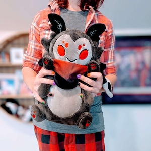 May include: A plush toy bat with a gray face, black wings, and red accents, held by a person. The bat has a red scarf and a gray body. The feet are black with red heart-shaped pads. The plush toy is designed in a cartoon style.