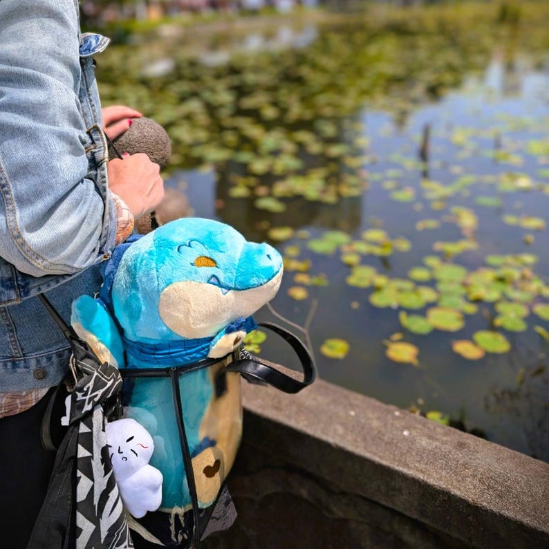 May include: A blue plush dinosaur toy with yellow eyes and a blue scarf is visible in a black and white patterned bag. A small white plush ghost toy is also in the bag. The background shows a pond with lily pads.