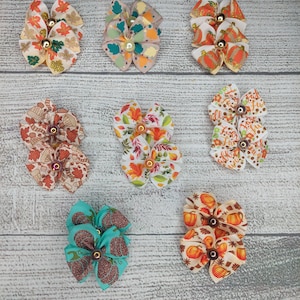 May include: Nine fall-themed hair bows with various patterns, including leaves, pumpkins, and acorns. The bows are made of fabric and have a gold center.