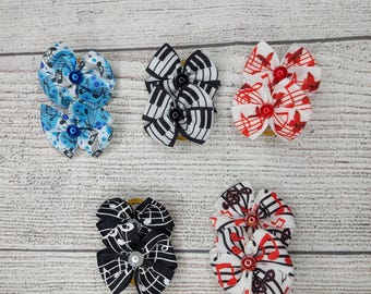 May include: Five decorative hair bows in various designs. The bows feature musical notes, piano keys, and other music-themed patterns in blue, black, and red. Each bow is centered with a decorative button or pearl.