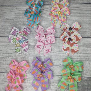 May include: A collection of colorful Easter-themed hair bows. The bows feature various patterns, including carrots, Easter eggs, and bunnies. The bows are in shades of pink, blue, green, and purple. Each bow has a decorative center.