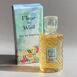 Può includere: Un piccolo flacone di profumo trasparente in vetro di Fleur de Weil Eau de Toilette. Il flacone è circondato da una scatola blu chiaro con un motivo floreale e il testo "Fleur de Weil Eau de Toilette" stampato su di essa. La scatola ha anche un piccolo logo "W" con il testo "Parfums Weil Paris" stampato sotto.