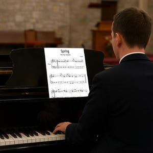 May include: A person playing a black grand piano. Sheet music titled "Spring" from "The Four Seasons" is on the music stand. The piano keys are white and black. The background is a church interior.