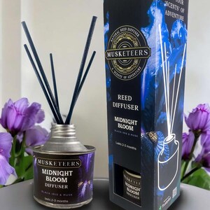 May include: A silver metal reed diffuser bottle with black reeds and a black and blue label that says "Musketeers Midnight Bloom Diffuser Black Iris & Musk Lasts 2-3 months". The bottle is sitting on a grey surface next to a box with the same design.