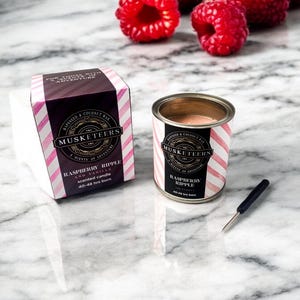 May include: A tin of "Raspberry Ripple" scented candle and its box, with pink and white striped design, are on a marble surface. Fresh raspberries are scattered around. The candle tin and box both feature the text "Musketeers".