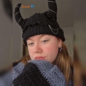 May include: A black crocheted beanie with long, black crocheted horns. The beanie has a silver chain detail and the horns have silver thread woven into them.