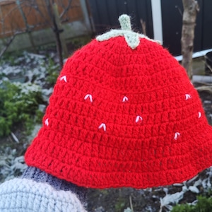 May include: Red crocheted bucket hat shaped like a strawberry with white embroidered details. The hat has a green crocheted top.