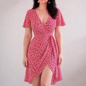 Op de afbeelding: Een rode wikkeljurk met een wit bloemenpatroon. De jurk heeft korte mouwen, een V-hals en een tailleband. De rok heeft een asymmetrische zoom, die een deel van het been onthult. De jurk is gemaakt van een lichte stof.