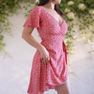 Puede incluir: Un vestido cruzado rojo con un estampado floral blanco. El vestido presenta mangas cortas y con volantes y un cierre de lazo en la cintura. La tela cae hasta un dobladillo ligeramente asimétrico.