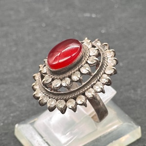 Peut inclure: Bague en argent ornée d'une grosse pierre rouge ovale au centre. La pierre est entourée de petites pierres claires et d'un filigrane argenté décoratif. La bague est présentée sur un présentoir transparent.