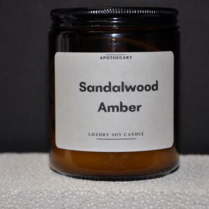 Pu&ograve; includere: Una candela in vetro marrone in un barattolo con un'etichetta bianca che recita "Apothecary Sandalwood Amber Luxury Soy Candle".