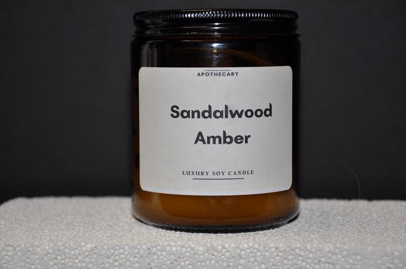 Pu&ograve; includere: Una candela in vetro marrone in un barattolo con un'etichetta bianca che recita "Apothecary Sandalwood Amber Luxury Soy Candle".