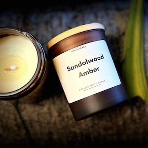Pu&ograve; includere: Un barattolo di vetro marrone per candele con un coperchio in legno. L'etichetta recita "Apothecary Sandalwood Amber Luxury Soy Candle".