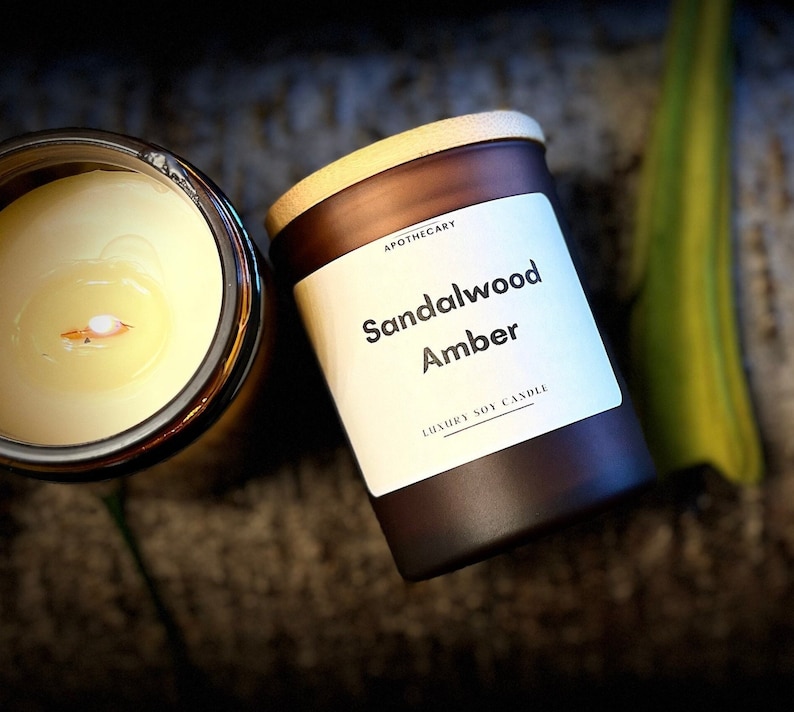 Pu&ograve; includere: Un barattolo di vetro marrone per candele con un coperchio in legno. L'etichetta recita "Apothecary Sandalwood Amber Luxury Soy Candle".