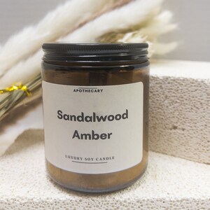 Pu&ograve; includere: Un barattolo di vetro marrone per candele con un coperchio nero. L'etichetta recita "APOTHECARY Sandalwood Amber LUXURY SOY CANDLE".