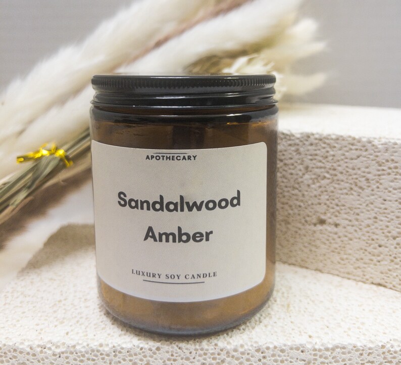 Pu&ograve; includere: Un barattolo di vetro marrone per candele con un coperchio nero. L'etichetta recita "APOTHECARY Sandalwood Amber LUXURY SOY CANDLE".