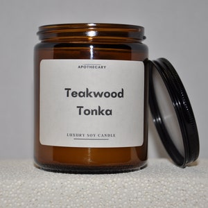 May include: A brown glass candle jar with a white label that reads "Apothecary Teakwood Tonka Luxury Soy Candle". The lid is black and is sitting next to the jar.