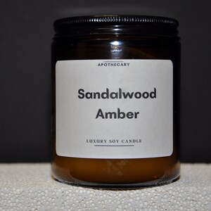 Pu&ograve; includere: Un barattolo di vetro marrone con una candela e un'etichetta bianca su cui &egrave; scritto "Apothecary Sandalwood Amber Luxury Soy Candle".