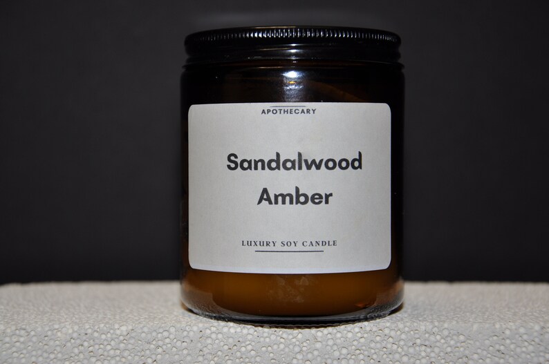 Pu&ograve; includere: Un barattolo di vetro marrone con una candela e un'etichetta bianca su cui &egrave; scritto "Apothecary Sandalwood Amber Luxury Soy Candle".